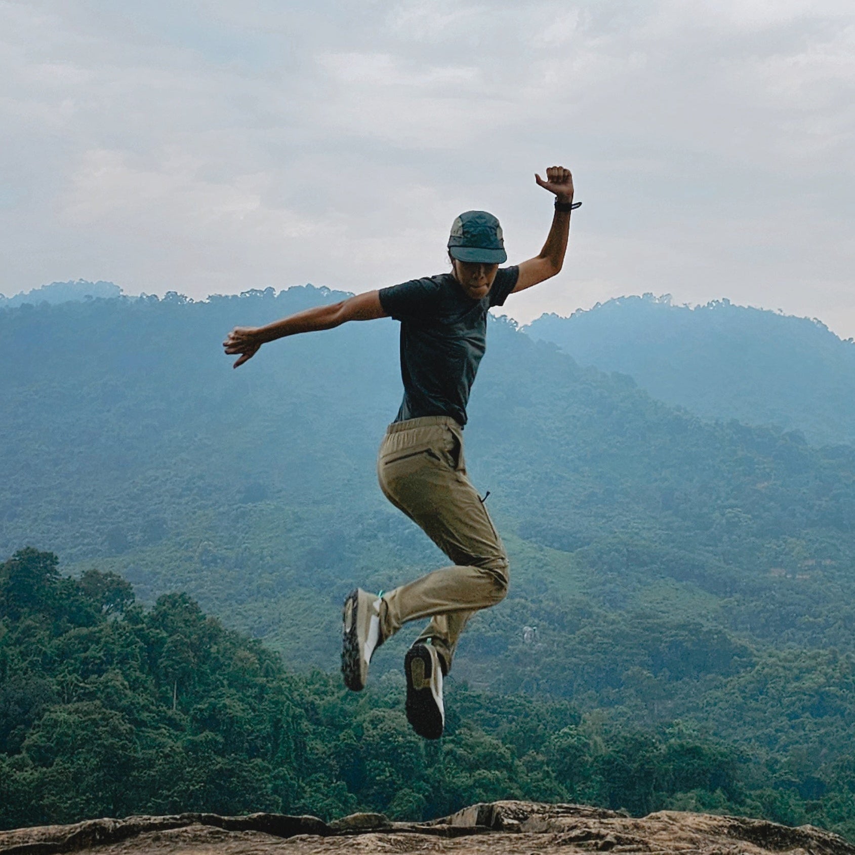 Person jumping on a rocky outcrop with a mountainous landscape and cloudy sky in the background